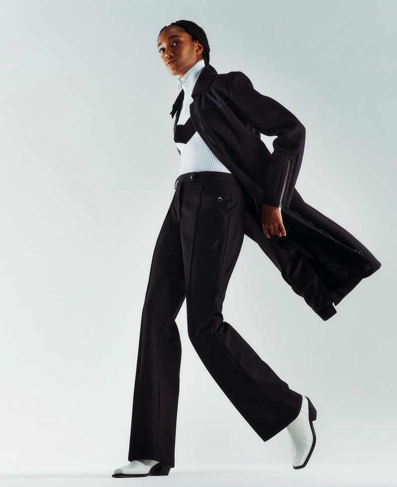 A dark-skinned fashion model strides confidently in black pants, top, and jacket paired with striking white heeled boots against a white backdrop.