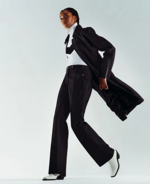 A dark-skinned fashion model strides confidently in black pants, top, and jacket paired with striking white heeled boots against a white backdrop.