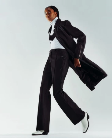 A dark-skinned fashion model strides confidently in black pants, top, and jacket paired with striking white heeled boots against a white backdrop.