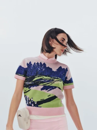 A woman with short brown hair poses in a pink sweater and skirt, wearing a golf-themed sweater and white belt bag against a white backdrop.