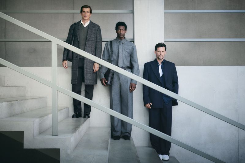 Three adult men in formal attire standing on a concrete staircase indoors, showcasing fashion with neutral color tones and modern architecture.