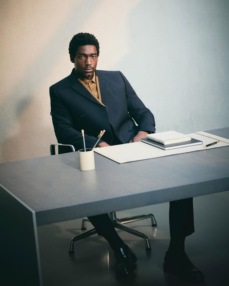 Portrait of a Black man in a business suit at an office desk with notebooks, pen, and pencil holder in front of a neutral backdrop.
