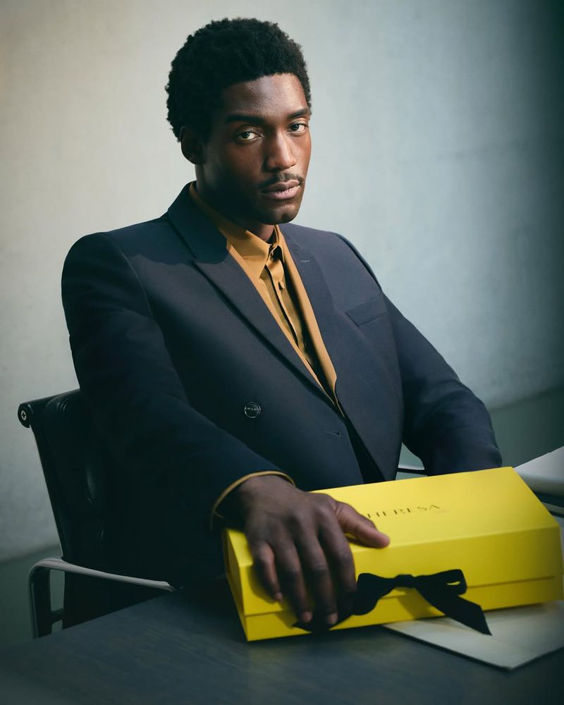 Portrait of a handsome African American man in a blue suit with a gold shirt, holding a yellow gift box with a black bow.