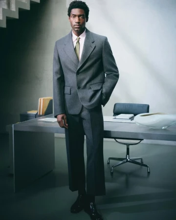 An African-American man in a grey suit stands in a modern office. A desk and chair are behind him, creating a professional and fashionable portrait.