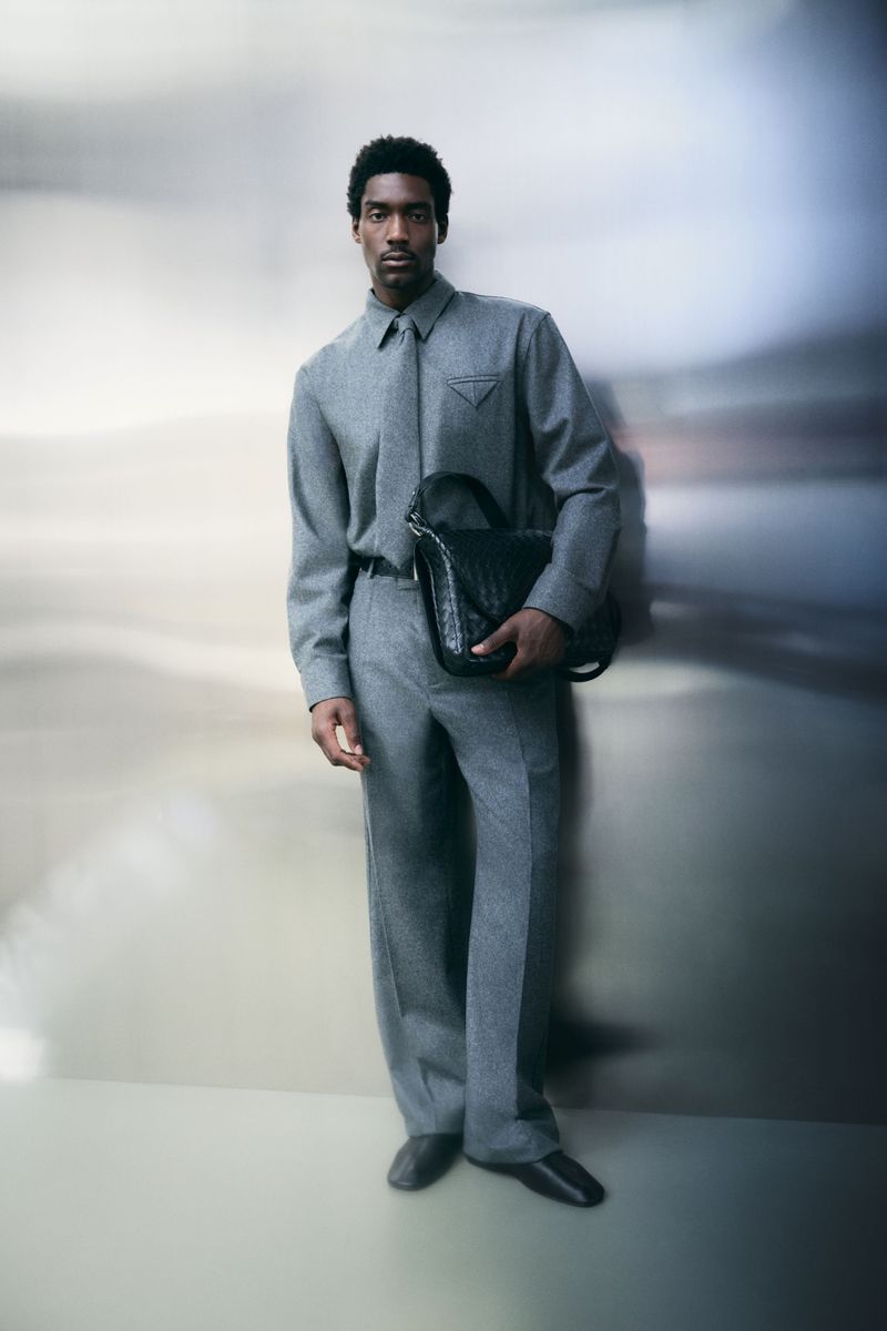 Full-length studio shot of a Black male model wearing a grey wool suit, carrying a black leather handbag, in front of a blurred backdrop.