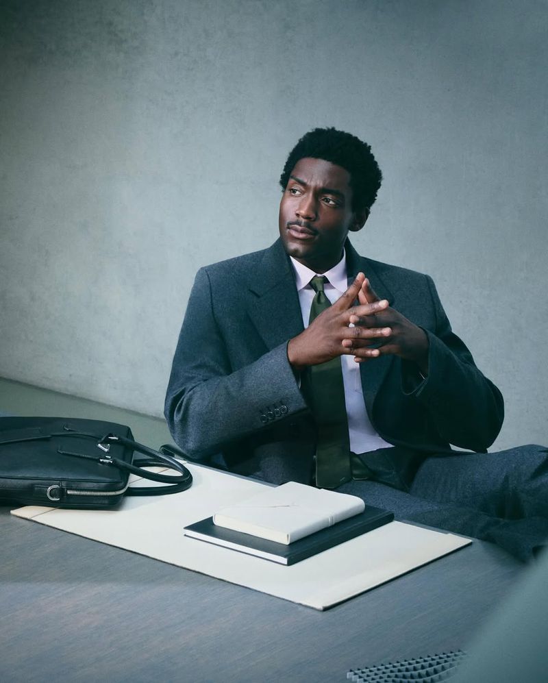 Portrait of a black male model in a suit sitting at a table with a briefcase and notebook in a photo studio with neutral grey wall.