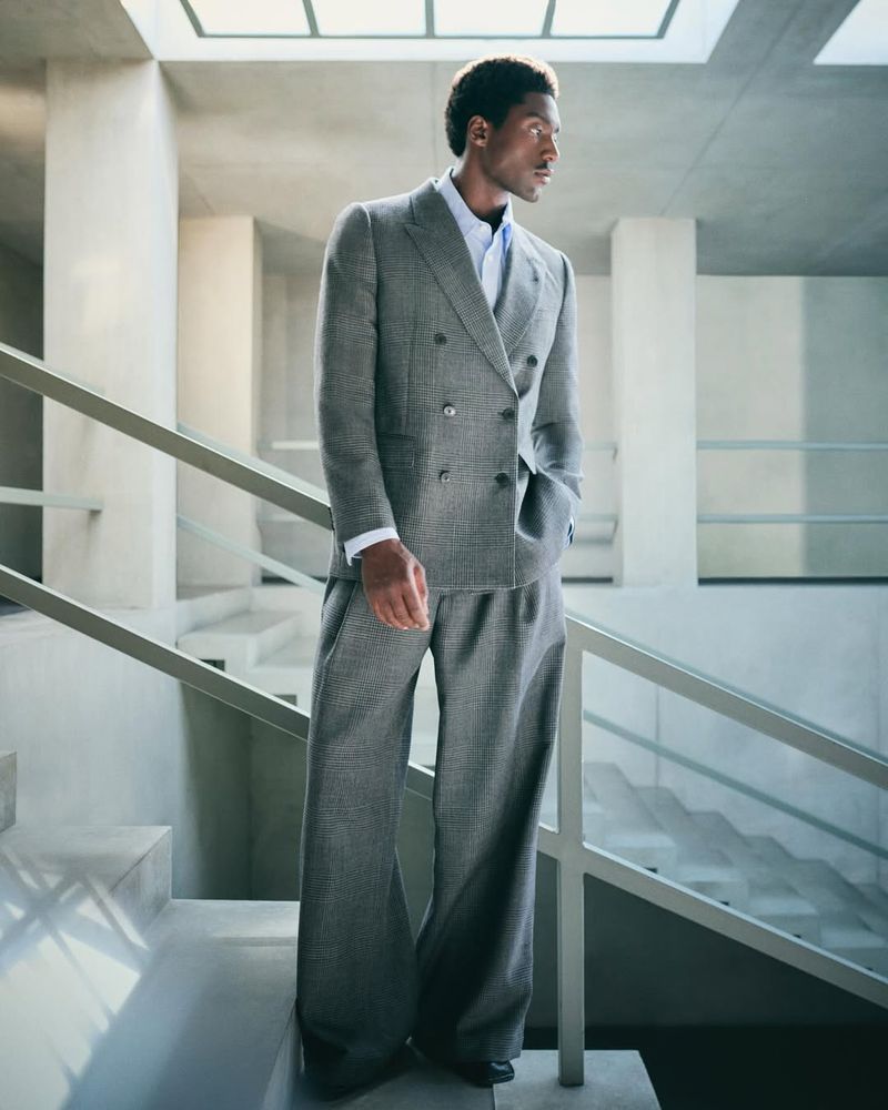 An African American man in a grey double-breasted suit stands on a staircase indoors with natural light from overhead windows.