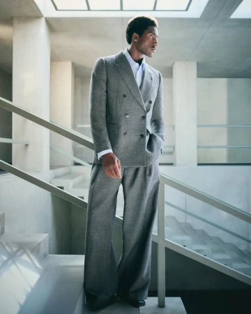 An African American man in a grey double-breasted suit stands on a staircase indoors with natural light from overhead windows.