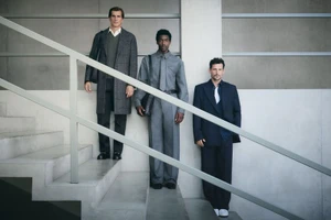Three male models in tailored menswear and suits standing on a modern concrete staircase behind a metal handrail.