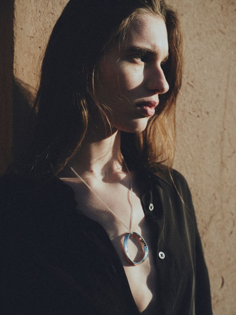 Woman wearing a black shirt and a silver circular pendant necklace against a peach-colored wall, partially in shadow and bright light.