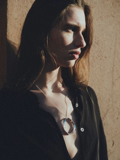 Woman wearing a black shirt and a silver circular pendant necklace against a peach-colored wall, partially in shadow and bright light.