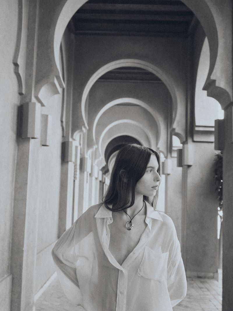 Woman in unbuttoned white shirt stands in an arched hallway, looking left with diffused lighting and monochrome coloration in the photo.