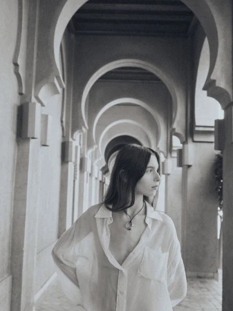 Woman in unbuttoned white shirt stands in an arched hallway, looking left with diffused lighting and monochrome coloration in the photo.