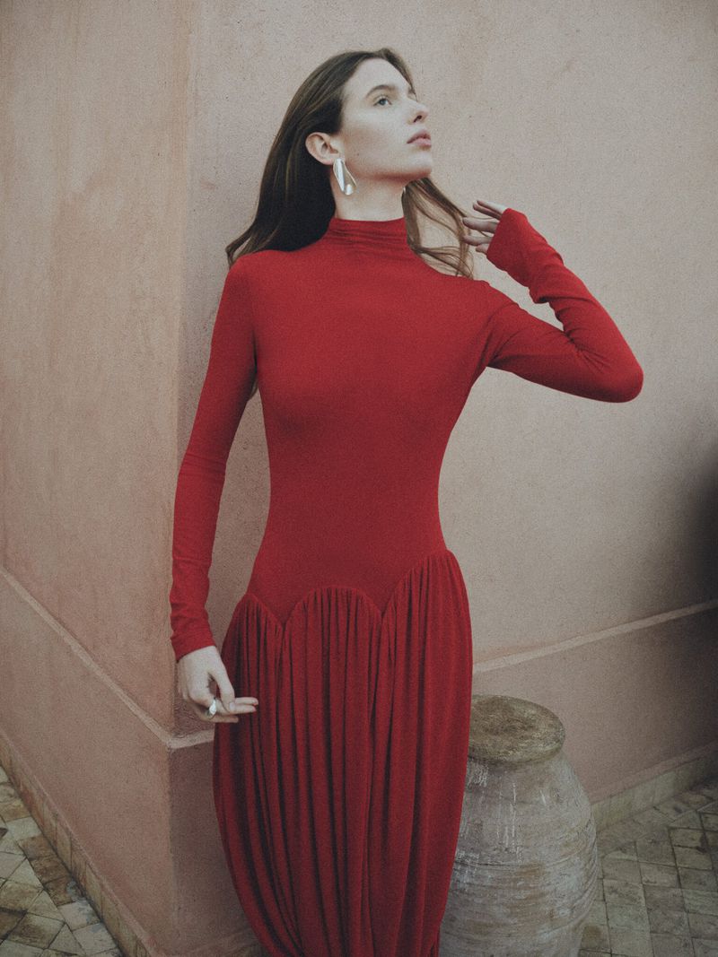 A young woman poses wearing a red dress with long sleeves next to a textured wall and ceramic pot. Fashion editorial photography.