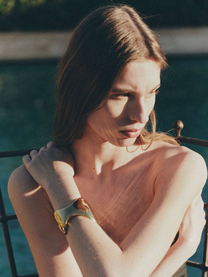 Model Esme Cornelius posing with arms crossed while wearing a sculptural gold cuff bracelet in warm outdoor lighting.