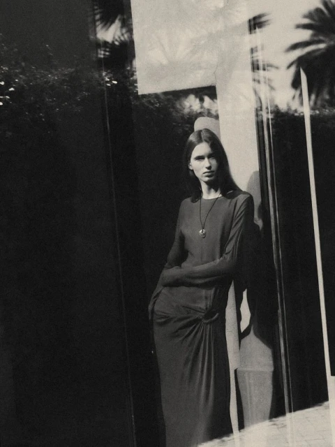 Monochrome portrait of a woman in a long dress leaning against a wall with reflected foliage and sunlight creating an elegant, moody scene.