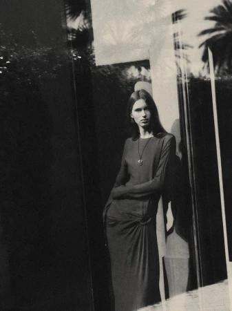 Monochrome portrait of a woman in a long dress leaning against a wall with reflected foliage and sunlight creating an elegant, moody scene.