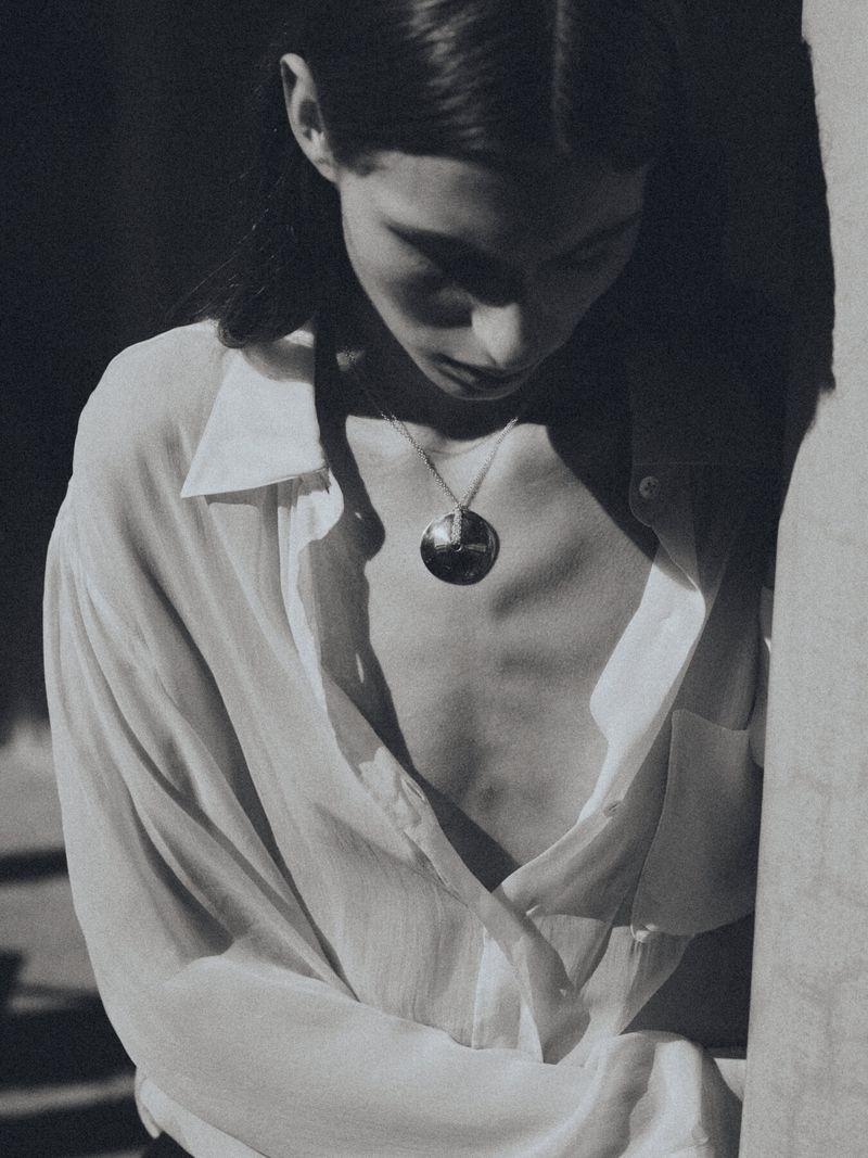 Monochromatic portrait of a young adult woman in an unbuttoned white shirt with a silver necklace, looking down in natural lighting.
