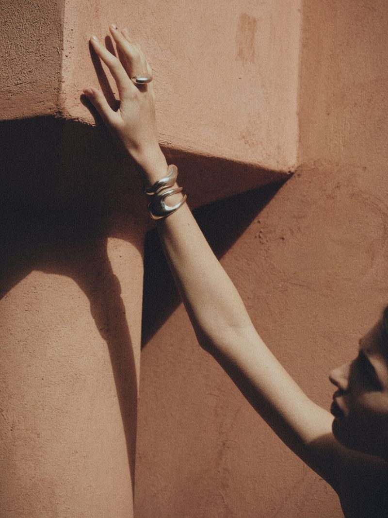 Model displaying silver jewellery including a ring and bracelets, posing against a textured terracotta wall, shot with natural lighting.