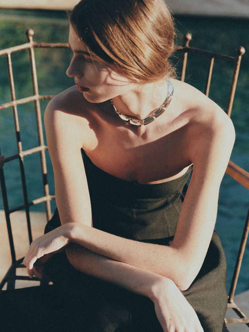 Woman with arms crossed wearing a silver necklace, seated outside on a sunny day, background shows a glimpse of dark green water.