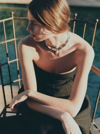 Woman with arms crossed wearing a silver necklace, seated outside on a sunny day, background shows a glimpse of dark green water.