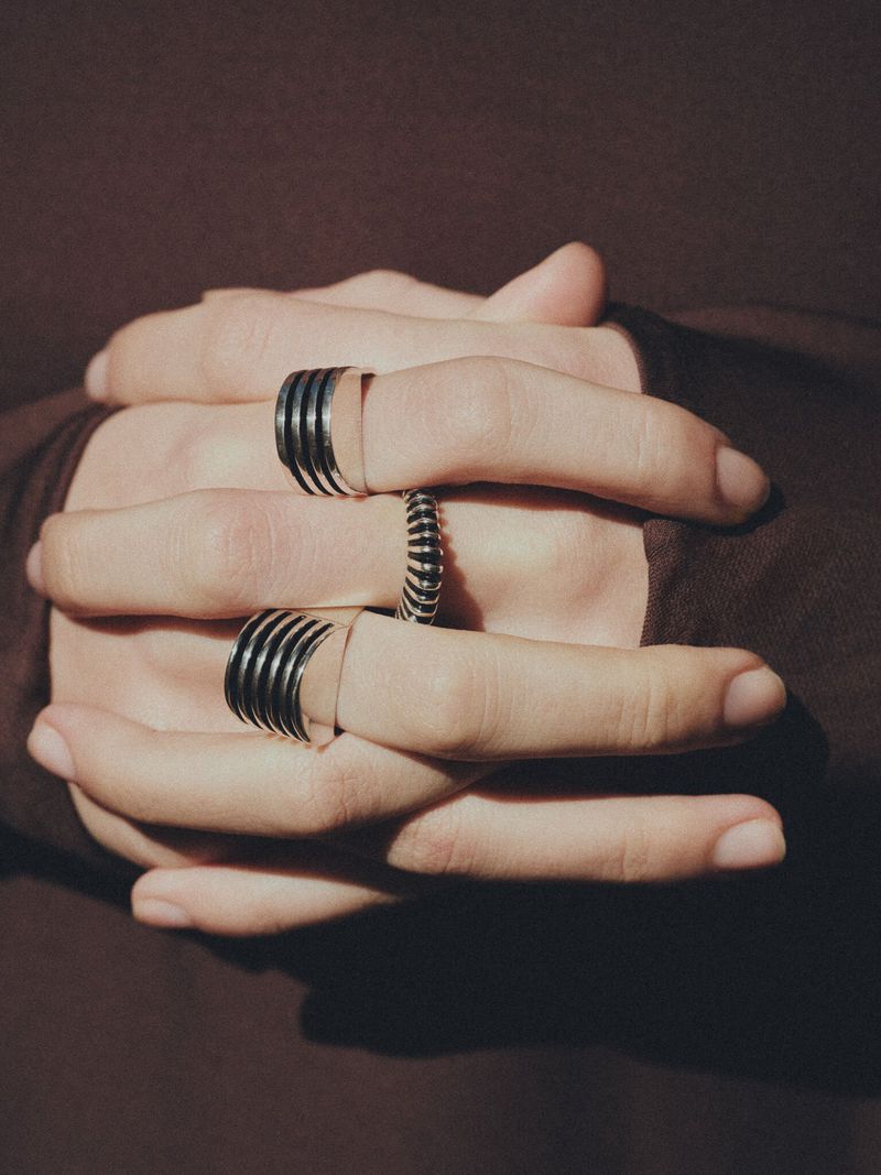 Close-up of a pair of hands folded together adorned with modern metal rings, set against a simple brown backdrop. Jewelry.