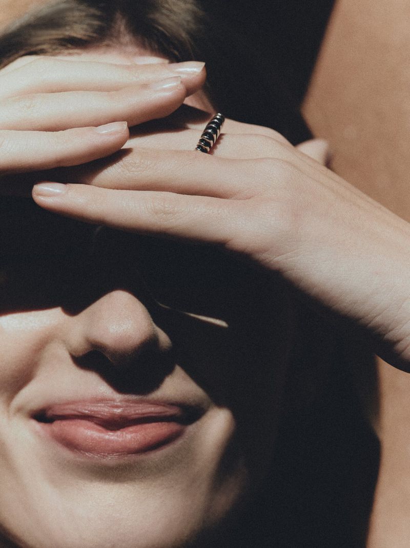 Close-up studio shot of a woman with her face partially covered by her hands, showcasing a ring on her finger in warm lighting.