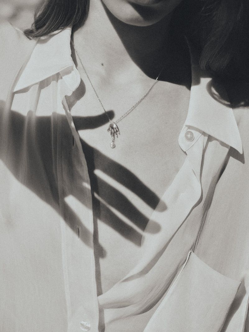 Close-up of a woman wearing a white collared shirt and a delicate pendant necklace, with shadows of a hand across her chest.