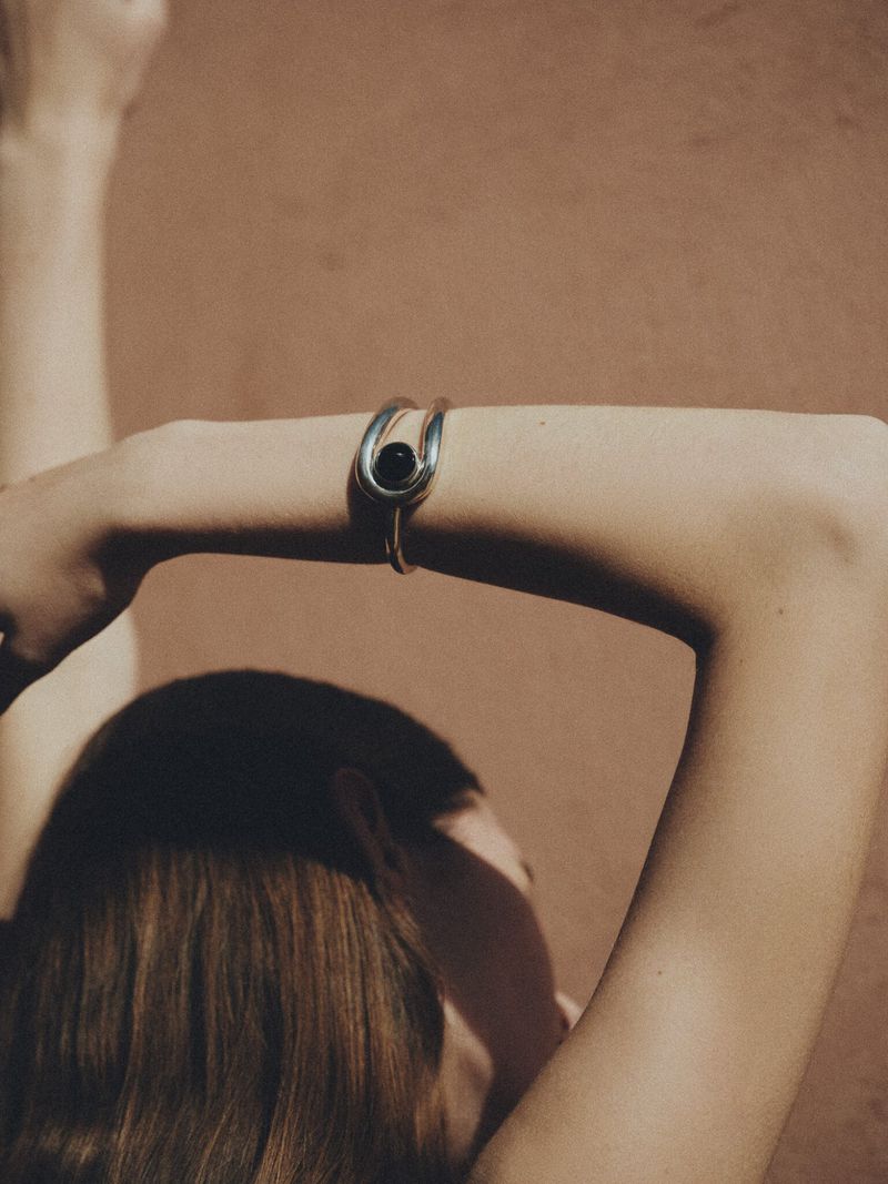 Woman with raised arms wearing a silver bracelet with a black bead on her wrist against a neutral tan background in a studio.