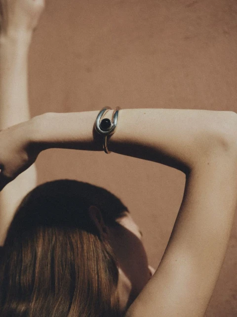 Woman with raised arms wearing a silver bracelet with a black bead on her wrist against a neutral tan background in a studio.