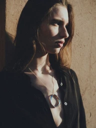 Model wearing a black shirt and silver pendant necklace standing against a textured wall in warm sunlight