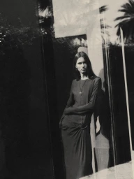 Model standing outdoors wearing a long dark dress and a pendant necklace, captured in a grainy monochrome photographic style.