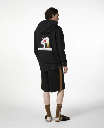 Man in black hooded sweatshirt with cartoon graphic and shorts against white background, fashion forward, studio shot.
