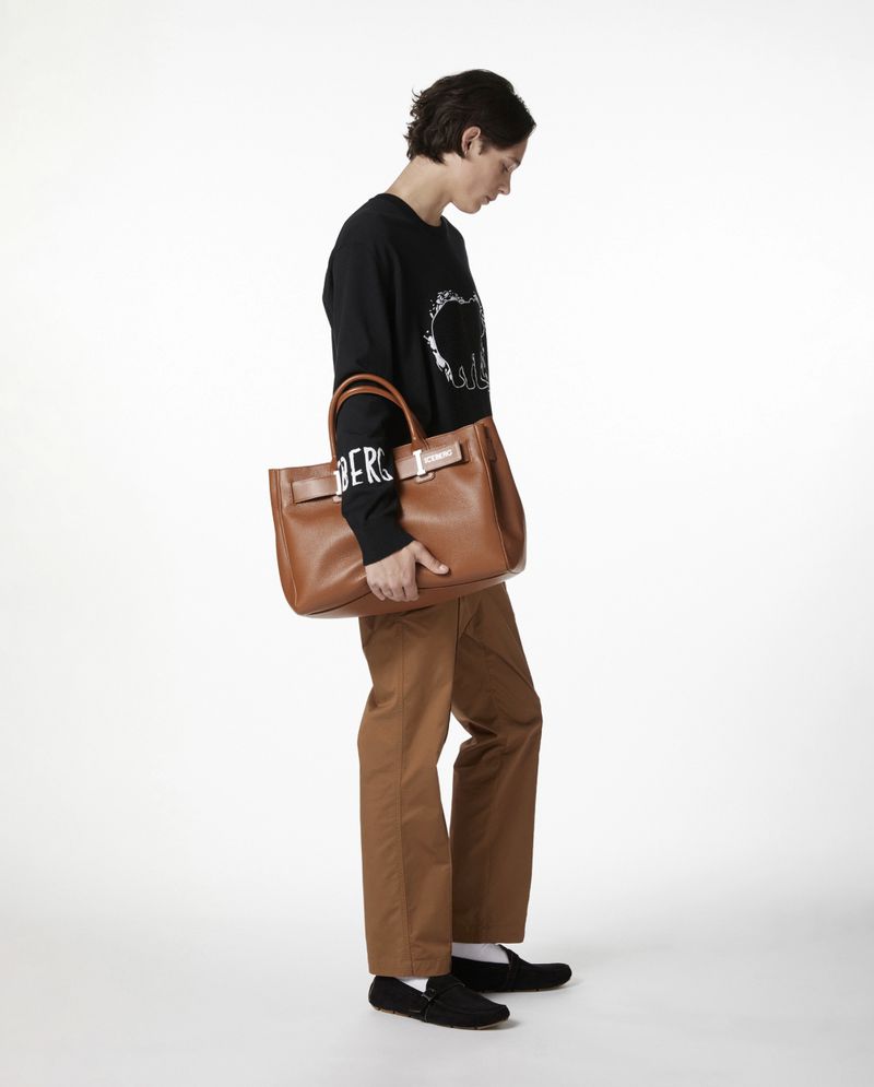 Adult wearing a black sweatshirt and brown pants standing with a brown leather tote bag in a bright studio against a white backdrop.