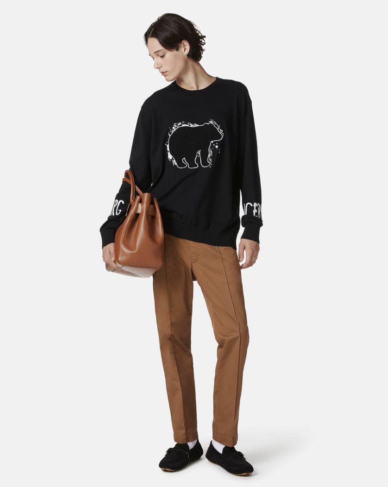 Full shot of a young adult in a black bear graphic sweatshirt, brown pants, and black flats holding a brown leather bag against a white background.