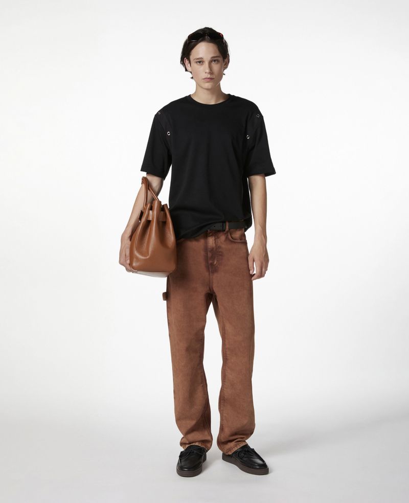 Male model wearing black t-shirt and brown denim pants, holding a brown leather bucket bag, posing in a white photo studio.