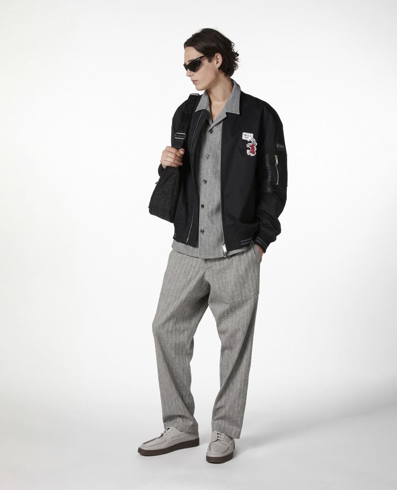 Adult male wearing a black bomber jacket, grey button shirt, and grey pants stands in a photo studio with a backpack.