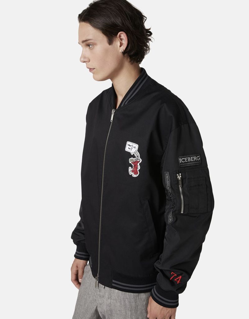 Adult male in a studio shot wearing a black Iceberg bomber jacket with a cartoon patch and grey pants, posing against a white backdrop.