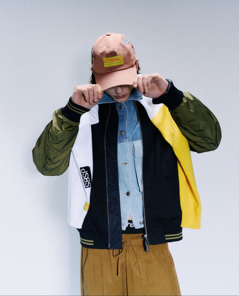 Adult in a color-blocked bomber jacket, brown corduroy pants, and baseball cap with hands near the collar and a light background.