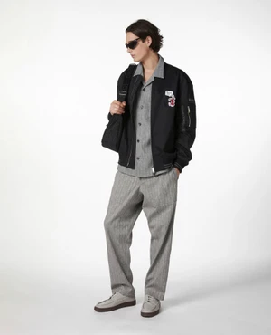 Full-length portrait of a male model wearing a black bomber jacket over grey patterned shirt and trousers against a white backdrop.