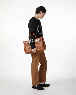 Model Etienne Maclaine in a black graphic sweatshirt and brown trousers holding a large brown leather tote bag against a white studio background.