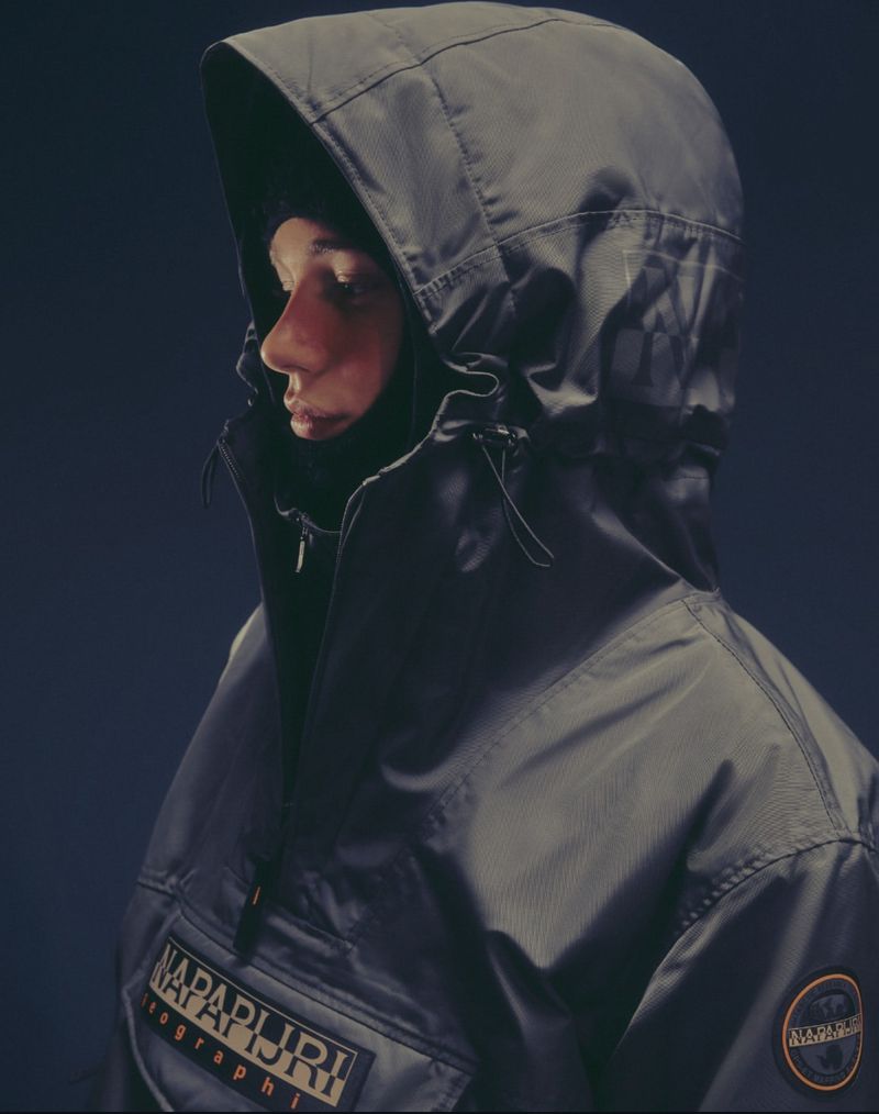 Person in a dark grey hooded jacket with a partial face covering against a dark background, outdoor or fashion apparel, cool lighting.