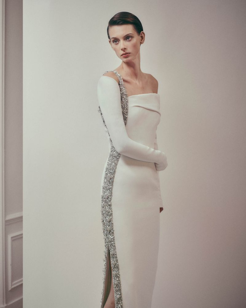 Elegant woman in a white one-shoulder gown with silver embellishments posing in a studio, showcasing modern high fashion.