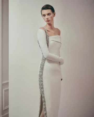 Elegant woman in a white one-shoulder gown with silver embellishments posing in a studio, showcasing modern high fashion.