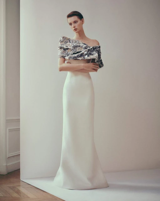 A woman poses indoors wearing a fitted white skirt and silver sequined asymmetrical top, set against a bright, neutral background.