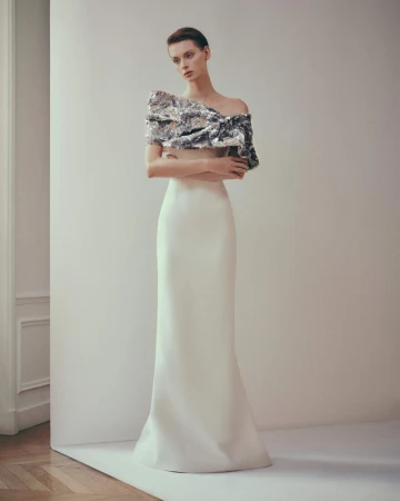 A woman poses indoors wearing a fitted white skirt and silver sequined asymmetrical top, set against a bright, neutral background.