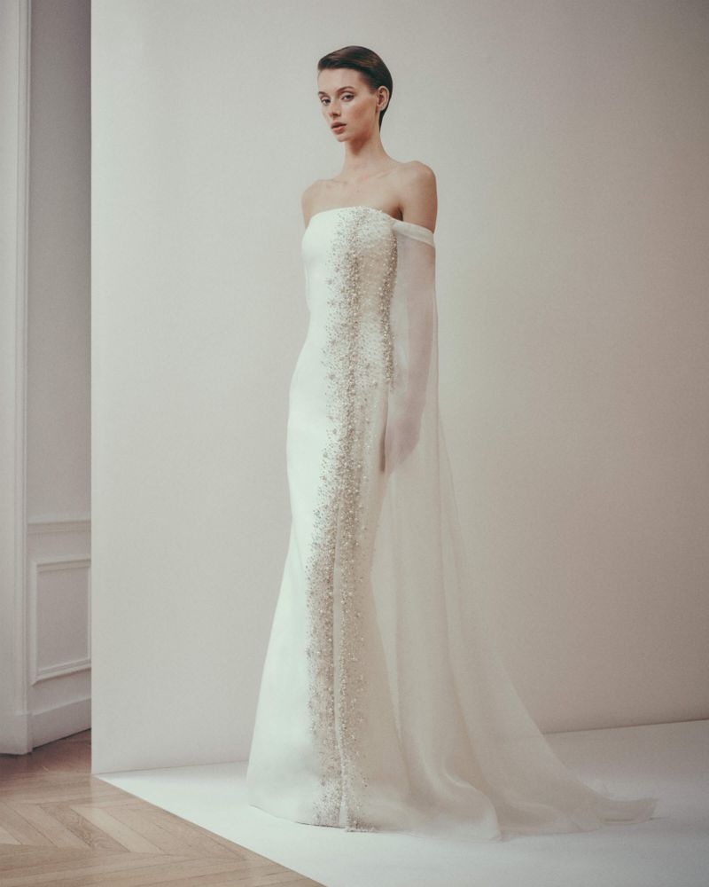 Elegant woman in strapless white gown with beaded embellishments and sheer cape, posing against a neutral background in a studio.