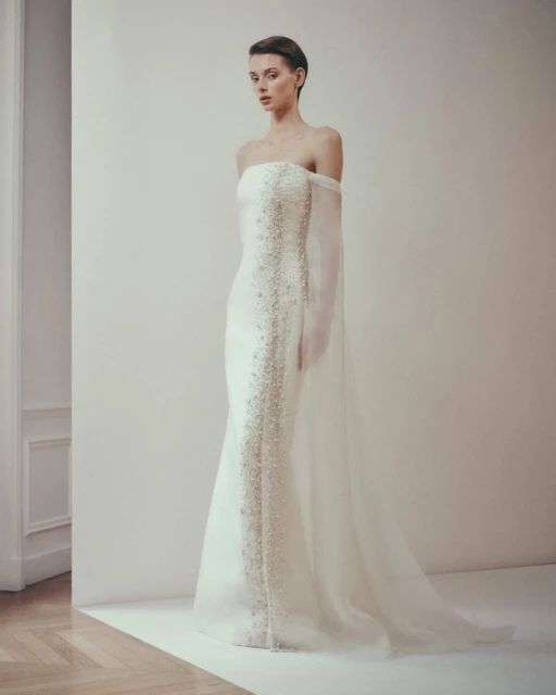 Elegant woman in strapless white gown with beaded embellishments and sheer cape, posing against a neutral background in a studio.