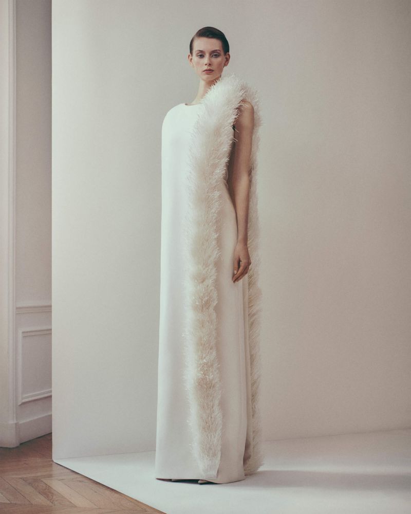 Elegant woman in a full-length white gown with fur-like trim posing against a neutral backdrop in a fashion editorial style shot.