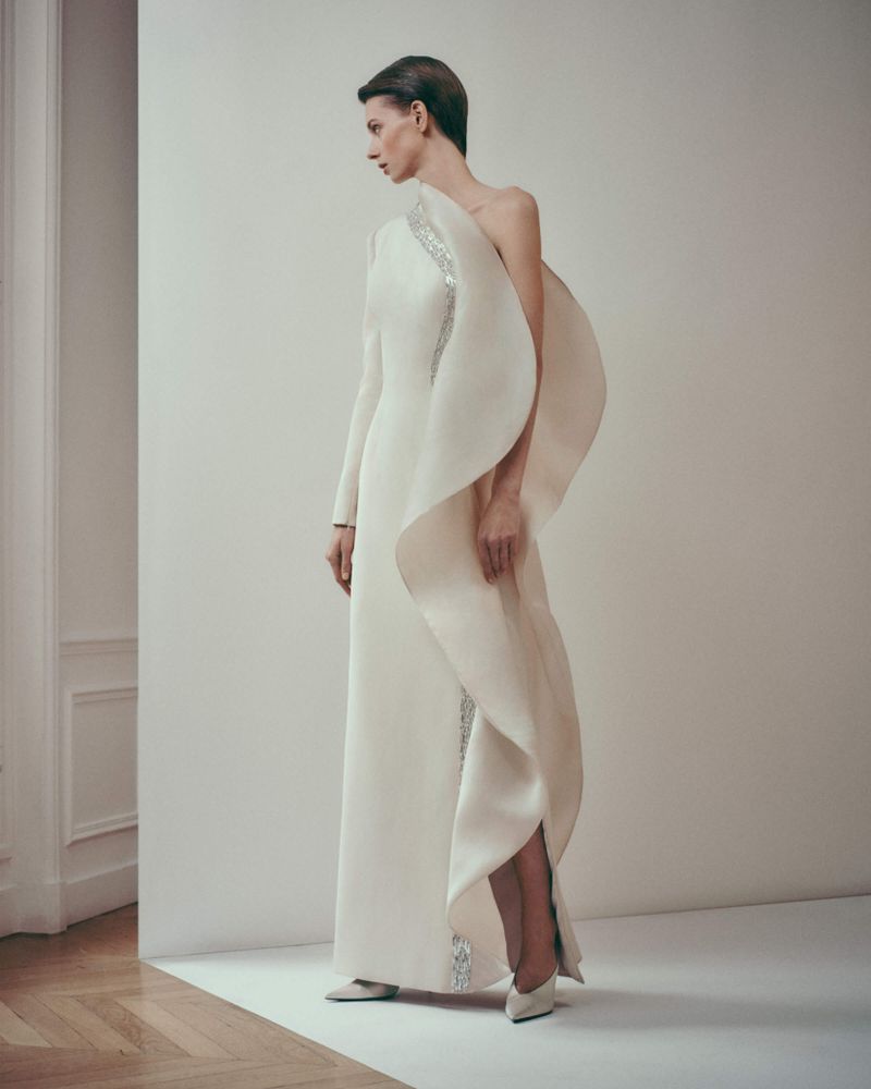 Elegant woman in a white gown poses in a studio. The gown has a one-shoulder design with draped fabric and embellishments.
